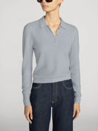 A model wearing a light gray textured knit polo sweater with a buttoned collar, paired with dark blue jeans, against a plain light gray studio background.