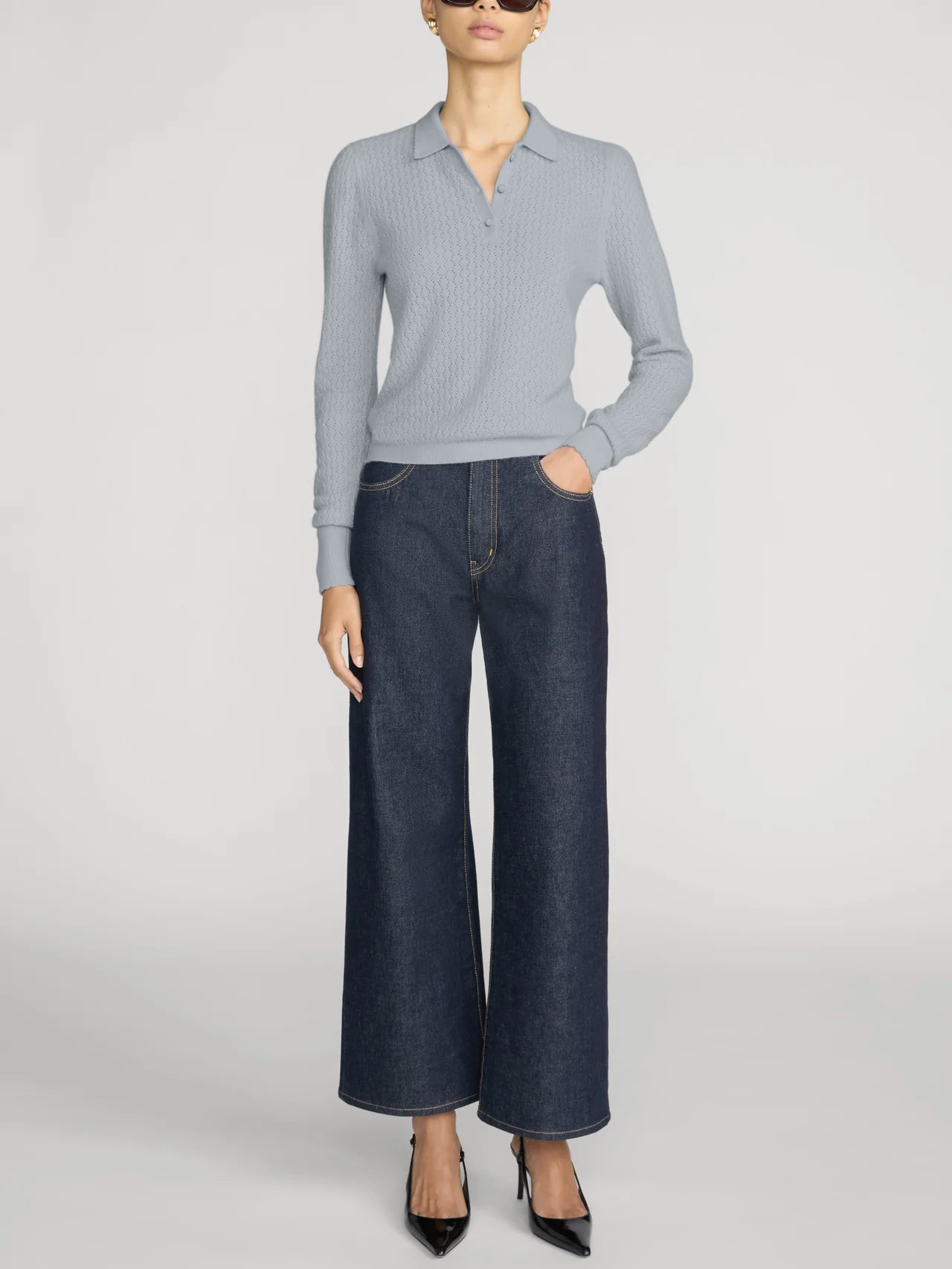 A model wearing a light blue ribbed knit polo shirt and dark blue wide-leg denim jeans, paired with black pointed-toe heels, against a light gray studio background.