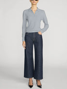 A model wearing a light blue ribbed knit polo shirt and dark blue wide-leg denim jeans, paired with black pointed-toe heels, against a light gray studio background.