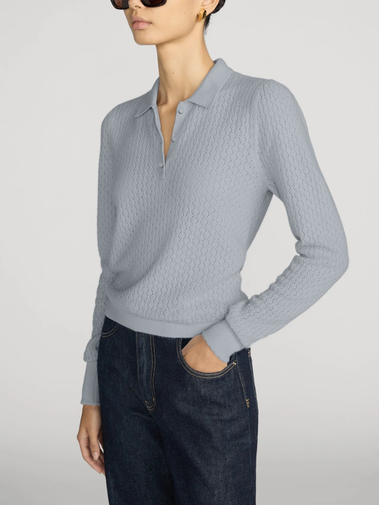 A woman wearing a light blue cable-knit polo sweater and dark blue jeans, shown from the side against a light gray studio background.