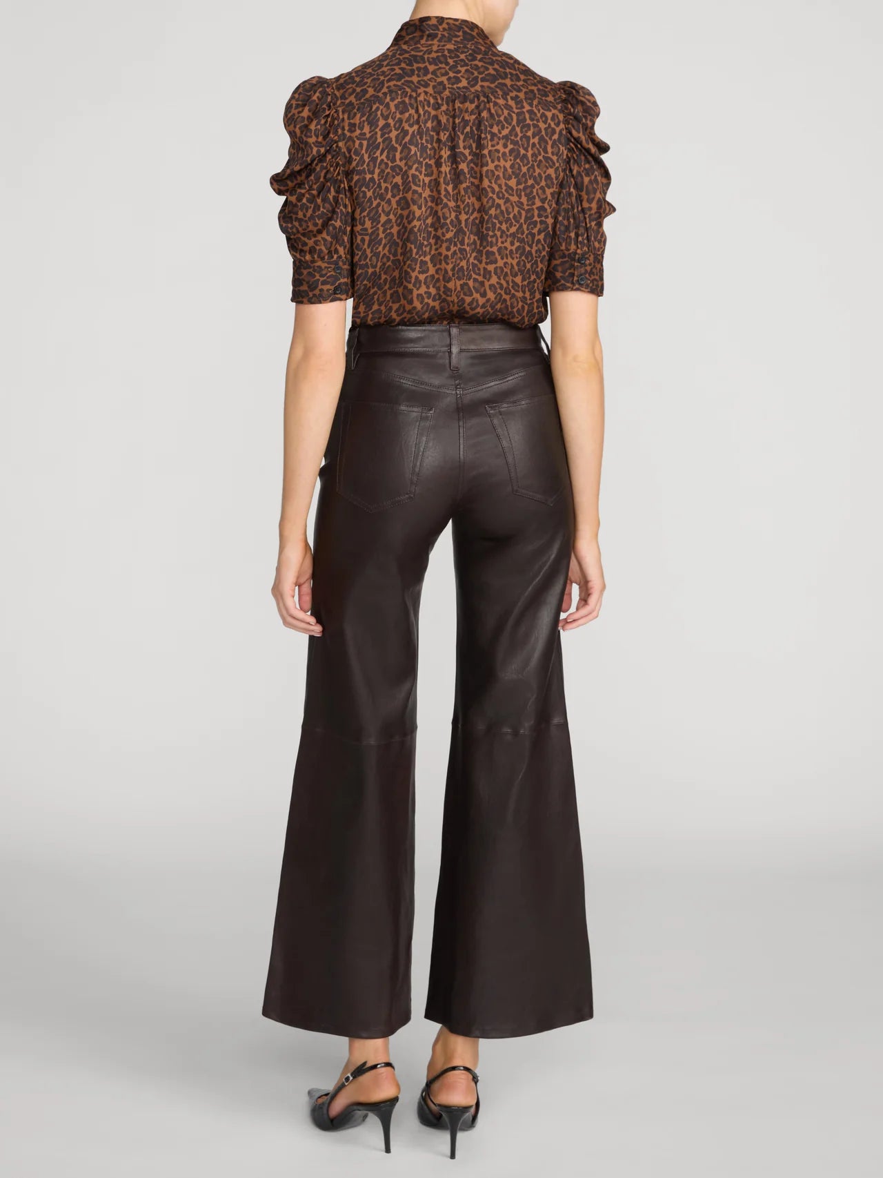 A model shown from the back wearing a brown and black leopard-print short-sleeve blouse with ruffled shoulders and dark brown wide-leg faux-leather pants, paired with black heels, against a light gray studio background.