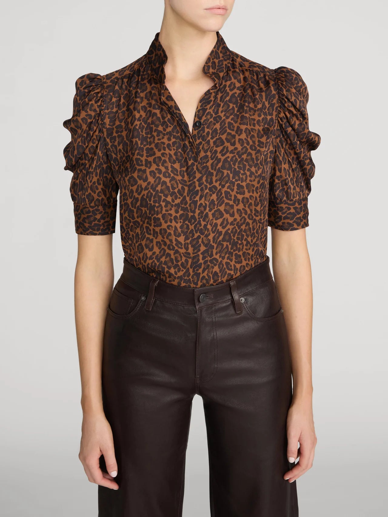 A model wearing a brown and black leopard-print blouse with dramatic ruffled puff sleeves, paired with dark brown faux-leather pants, against a light gray studio background.