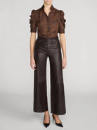 A model wearing a brown and black leopard-print short-sleeve blouse with ruffled sleeves, paired with dark brown wide-leg faux-leather pants and black pointed-toe heels, against a light gray studio background.