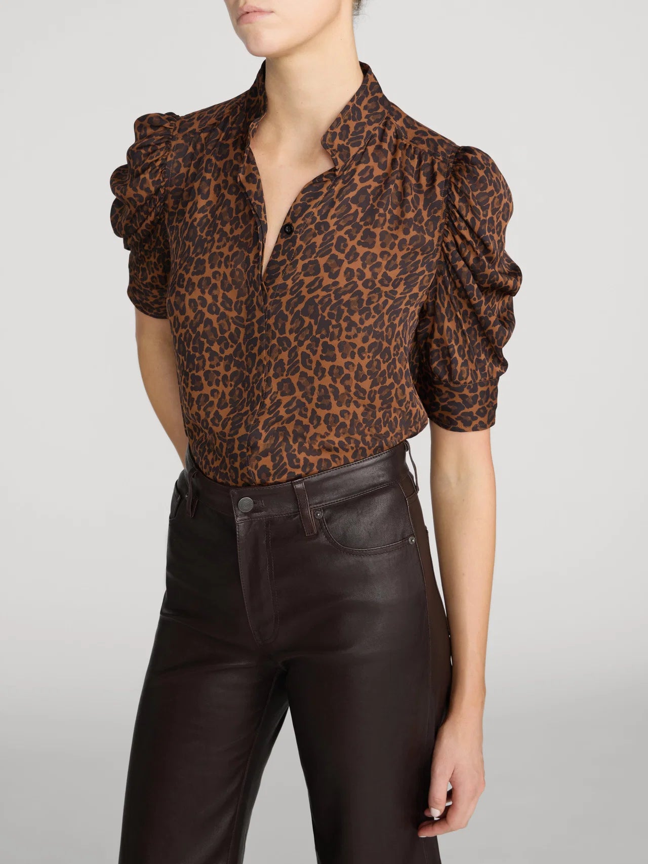 A model wearing a brown and black leopard-print blouse with dramatic puffed short sleeves and dark brown faux-leather pants, cropped from the chin down against a light gray studio background.