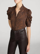 A model wearing a brown and black leopard-print blouse with dramatic puffed short sleeves and dark brown faux-leather pants, cropped from the chin down against a light gray studio background.