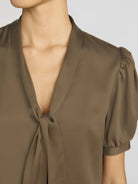 Close-up of a model wearing a taupe brown short-sleeve blouse with a deep V-neck and front tie detail, featuring puffed sleeves.