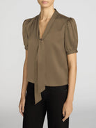 A woman wearing a taupe-brown short-sleeve blouse with puffed sleeves, a V-neckline, and a front tie detail, paired with black pants, against a light gray studio background.