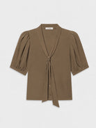 A taupe brown short-sleeve blouse with puffed sleeves, a wrap-style V-neckline, and a front tie detail, shown flat on a light gray background.