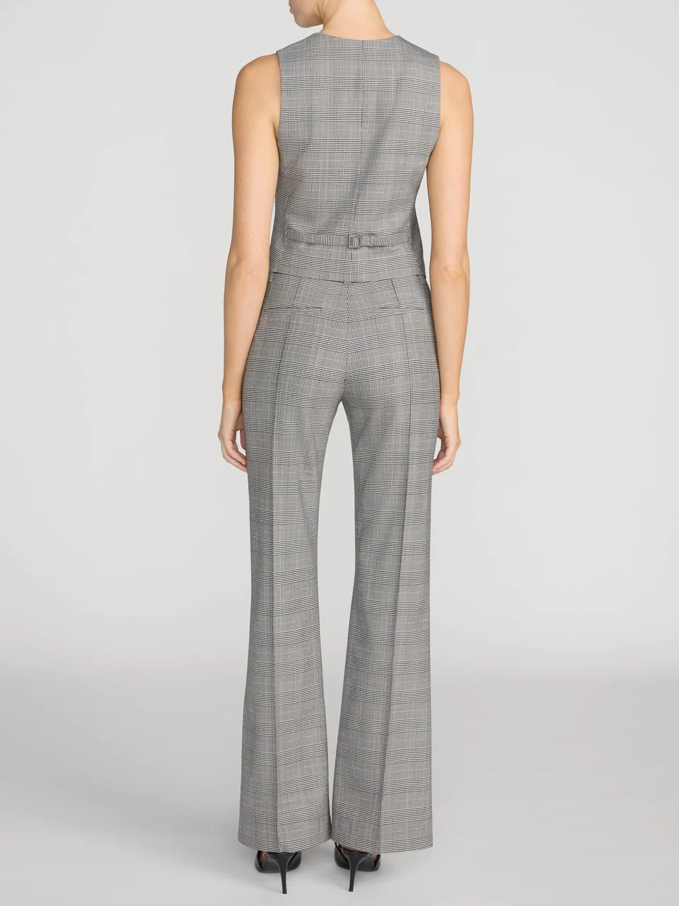 A model shown from the back wearing a gray sleeveless jumpsuit with a subtle plaid pattern, wide-leg pants, and a matching belt with a bow, set against a light gray studio background.