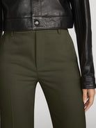 Close-up of a person wearing olive green tailored trousers with a black leather jacket cropped at the chest, against a light gray studio background.