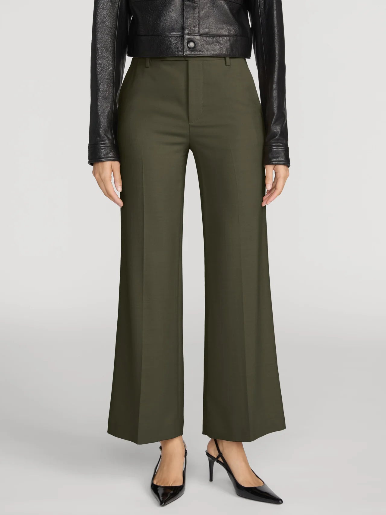 A model is shown from the waist down wearing olive green wide-leg trousers, a black leather jacket, and black pointed-toe heels against a light gray studio background.