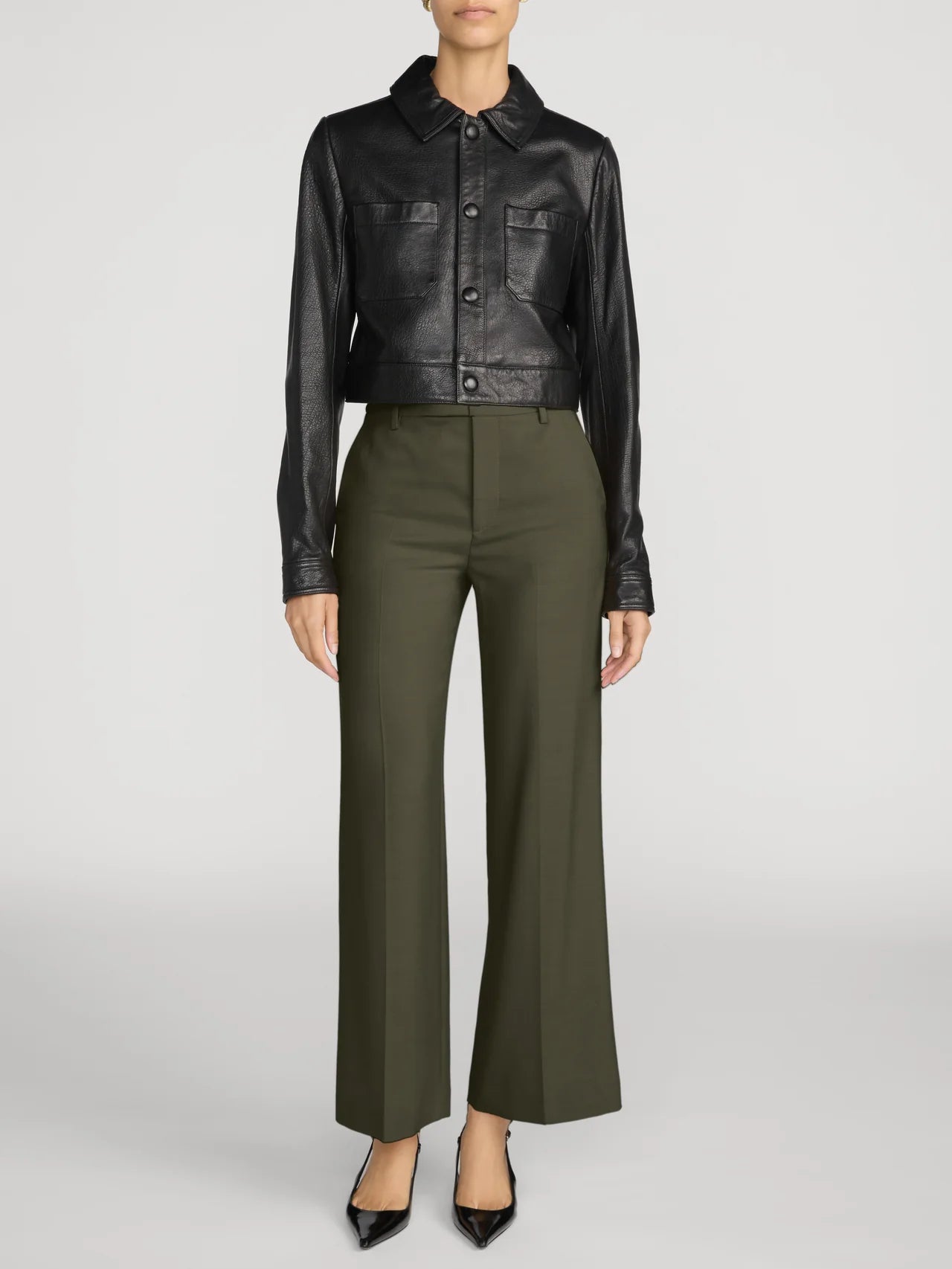 A model is shown from the neck down wearing a black leather cropped jacket and olive green wide-leg trousers, paired with black pointed-toe heels, against a light gray studio background.