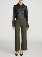 A model is shown from the neck down wearing a black leather cropped jacket and olive green wide-leg trousers, paired with black pointed-toe heels, against a light gray studio background.