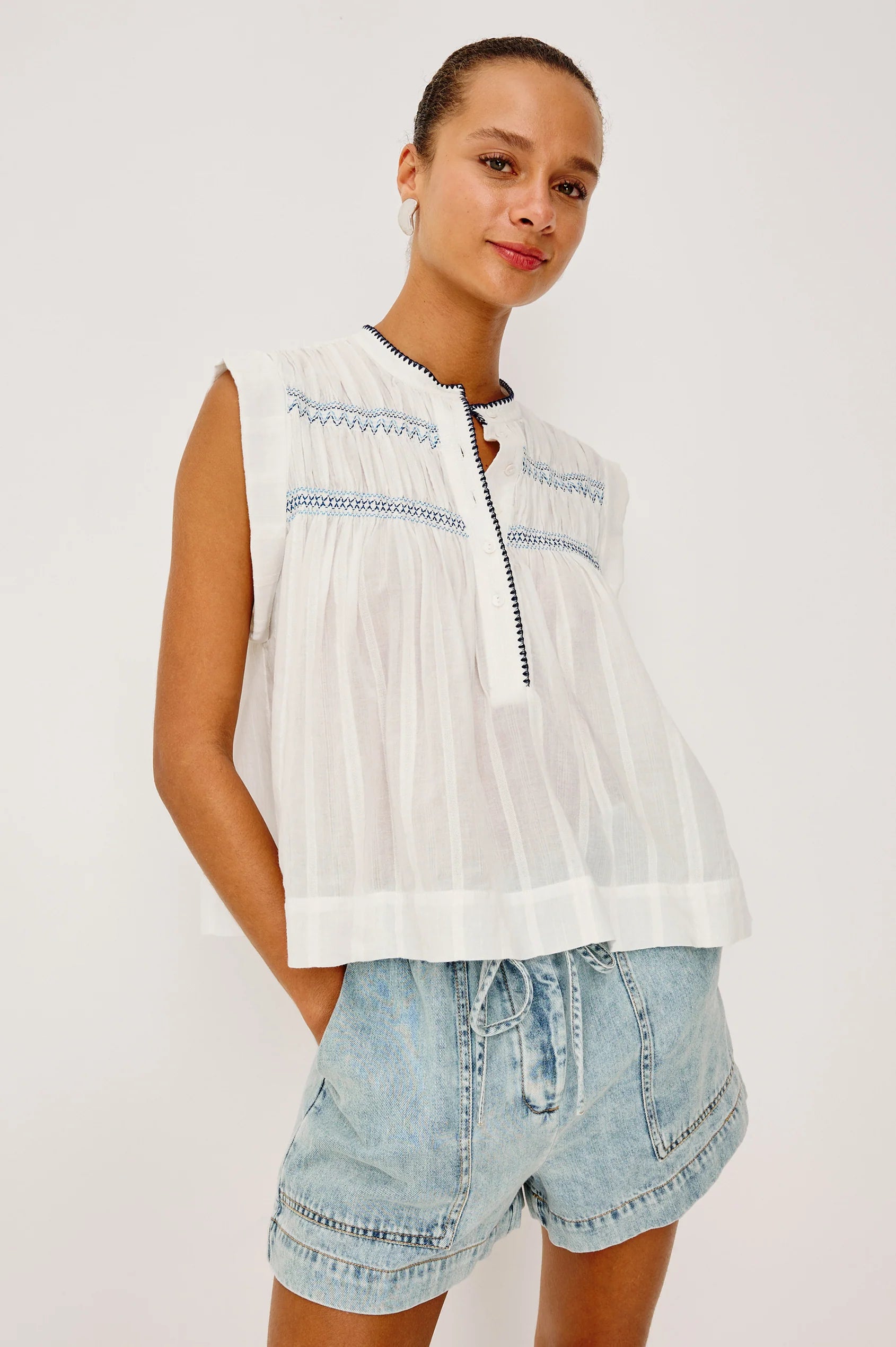 Woman wearing a sleeveless white top with blue embroidery and denim shorts on a plain background