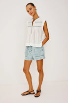 Woman wearing a sleeveless white top and denim shorts on a plain background