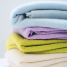 A stack of four folded knit fabric items in pastel colors, including light blue, lavender purple, chartreuse yellow-green, and cream white, photographed against a soft light background.