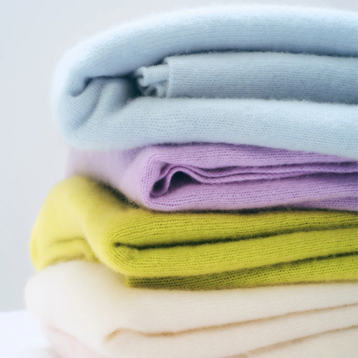 A stack of four folded knit fabric items in pastel colors, including light blue, lavender purple, chartreuse yellow-green, and cream white, photographed against a soft light background.