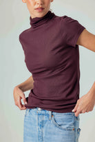 A woman wearing a short-sleeve, high-neck ribbed turtleneck top in a deep plum purple color, paired with light-wash blue jeans, against a solid light gray background.