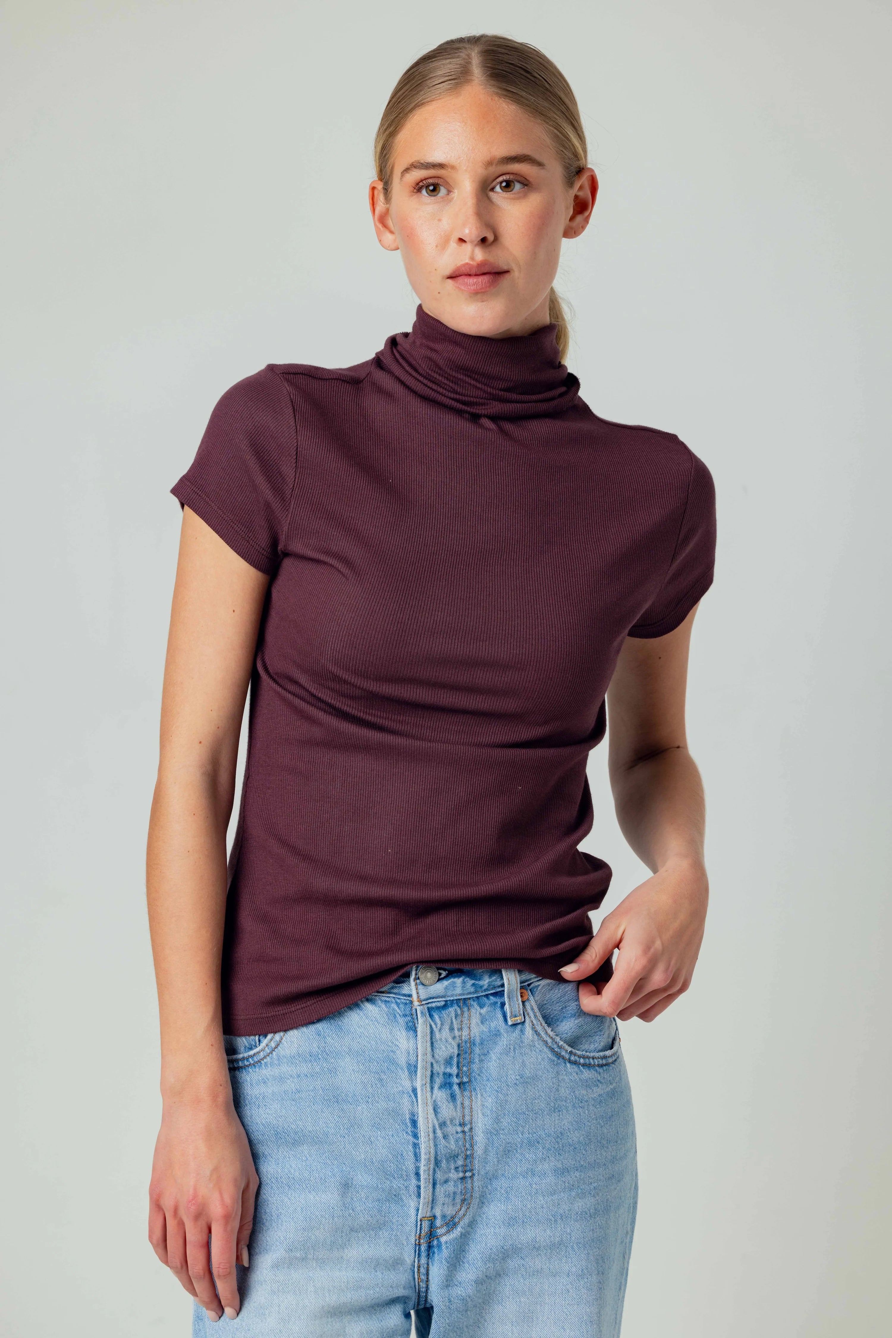 A woman modeling a short-sleeve, high-neck, ruched turtleneck top in a deep plum purple color, paired with light-wash blue jeans, against a plain light gray studio background.