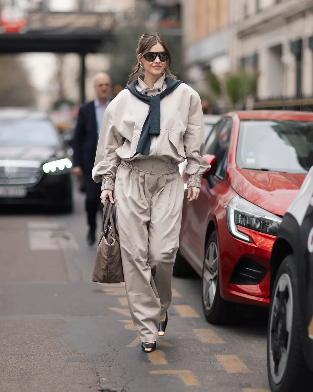Woman walking on a city street wearing a beige outfit with sunglasses and a scarf.