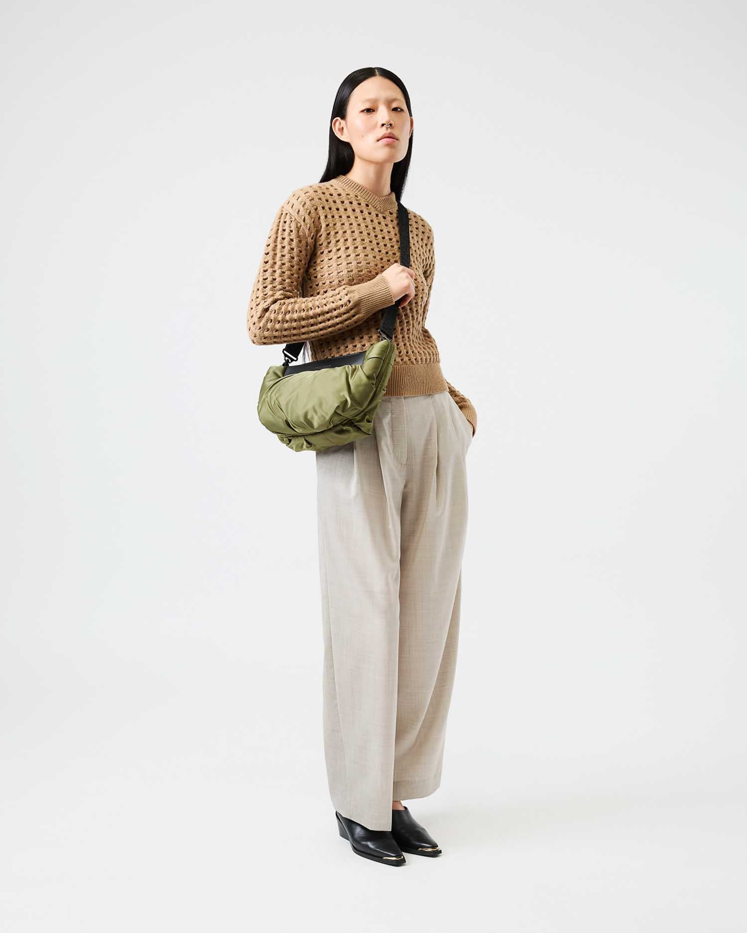 Woman wearing a brown knitted sweater and beige pants, holding a green bag on a white background