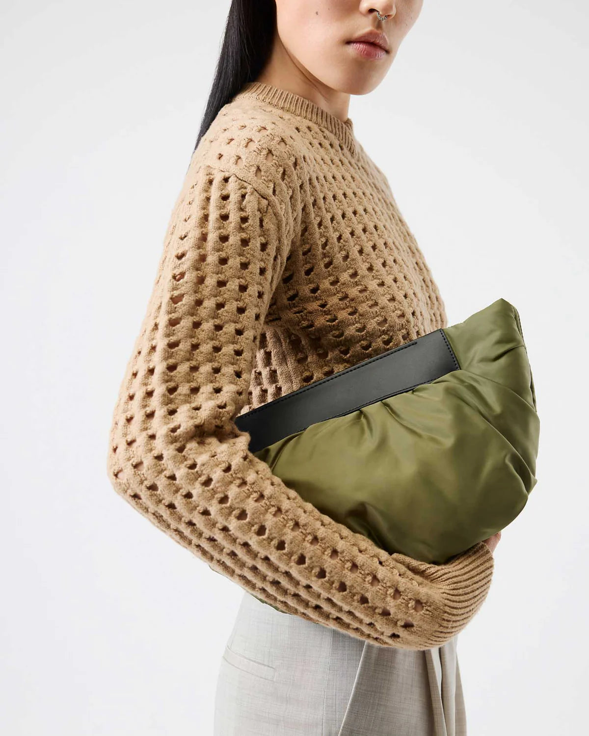 Person wearing a beige knit sweater holding an olive green bag against a white background