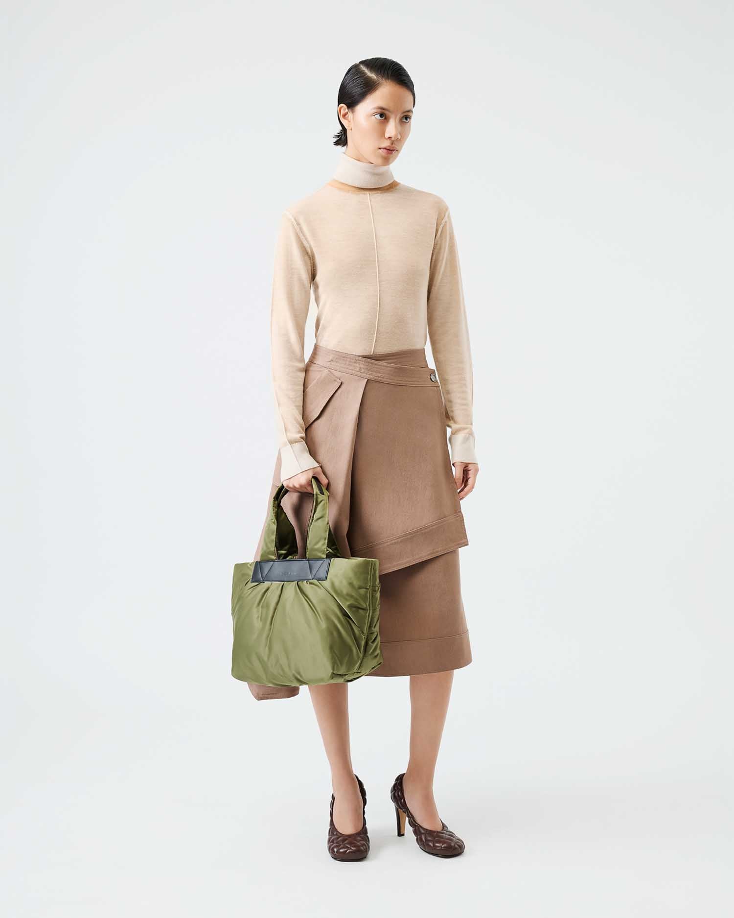 Woman holding a green bag wearing a beige sweater and brown skirt on a white background