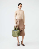 Woman holding a green bag wearing a beige sweater and brown skirt on a white background