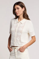 Woman wearing a white lace top and white pants against a plain background