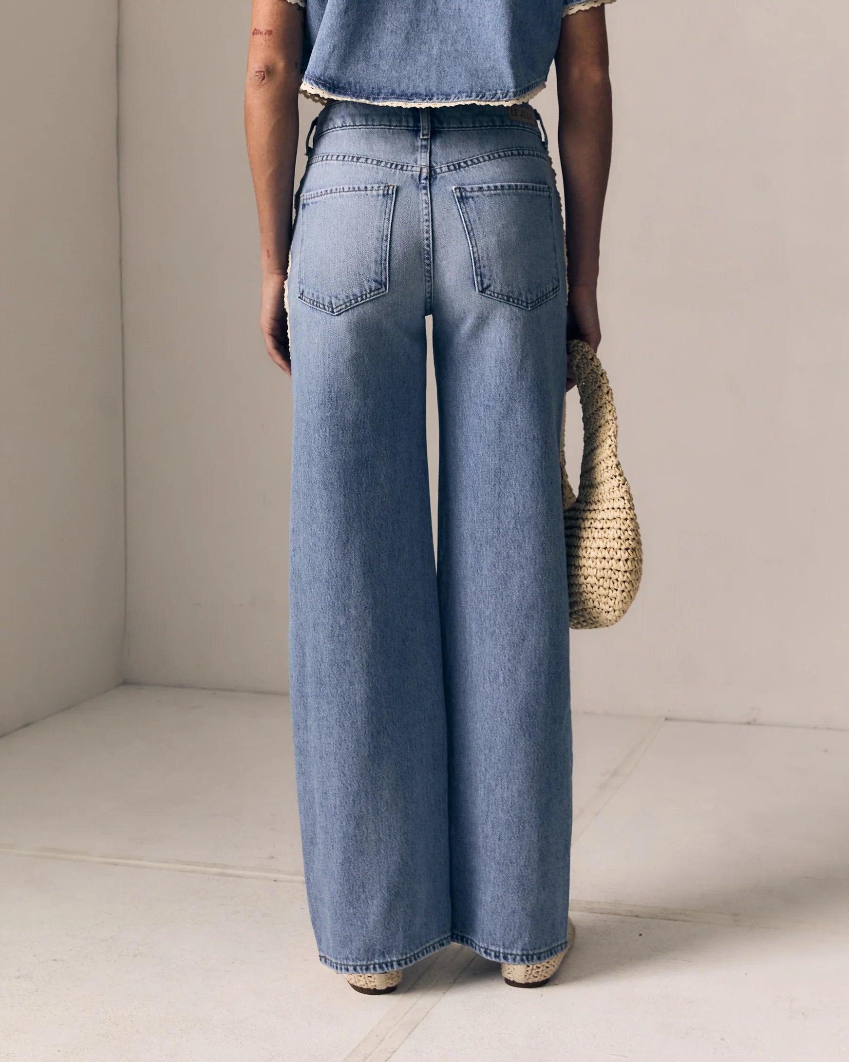 Person wearing light blue jeans holding a woven bag indoors.