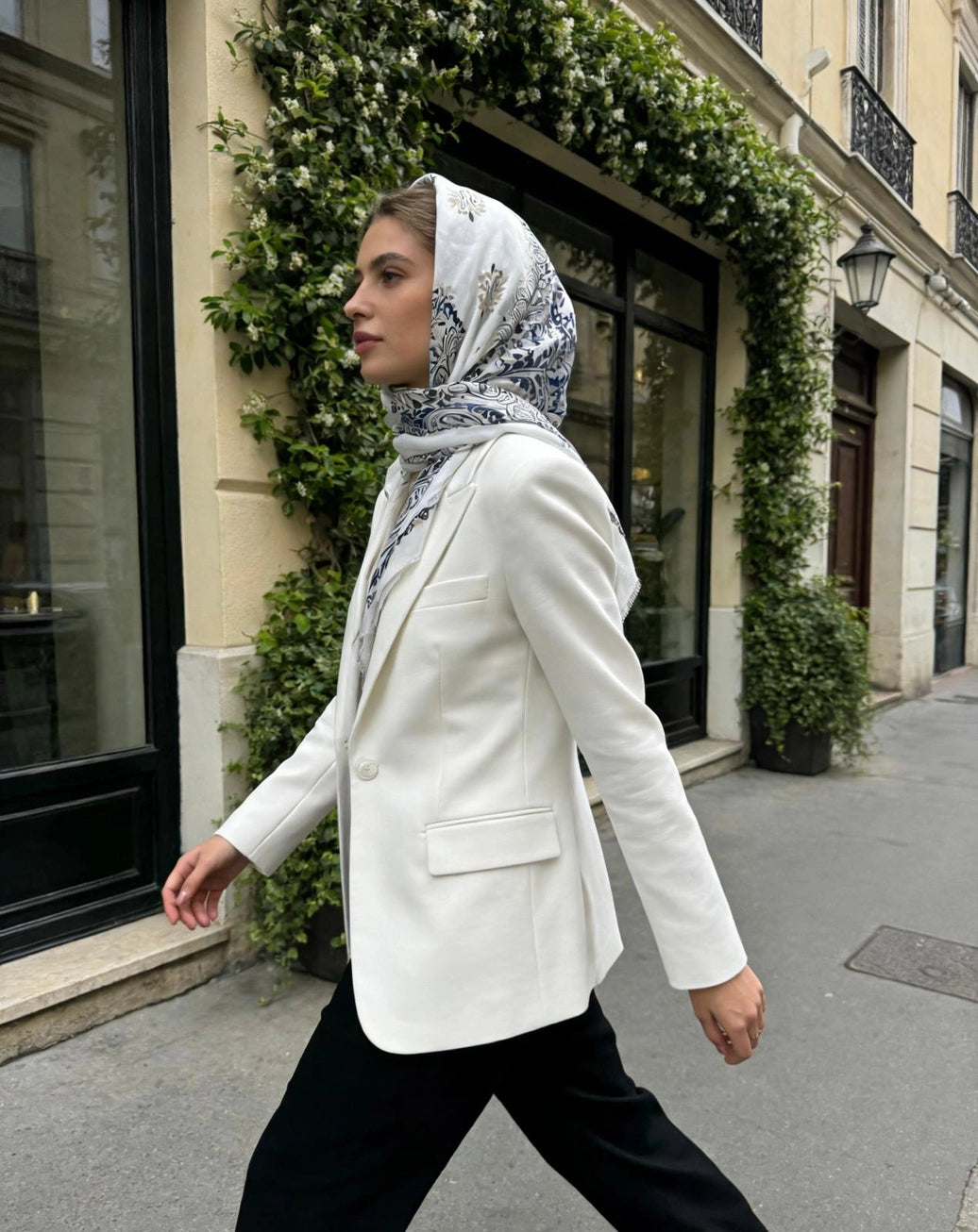 Woman wearing a white blazer and patterned scarf walking on a city street.