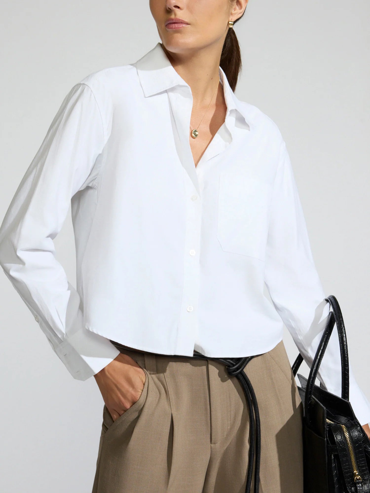 A woman wearing a white long-sleeve button-up shirt tucked into beige trousers, holding a black handbag against a light gray studio background.