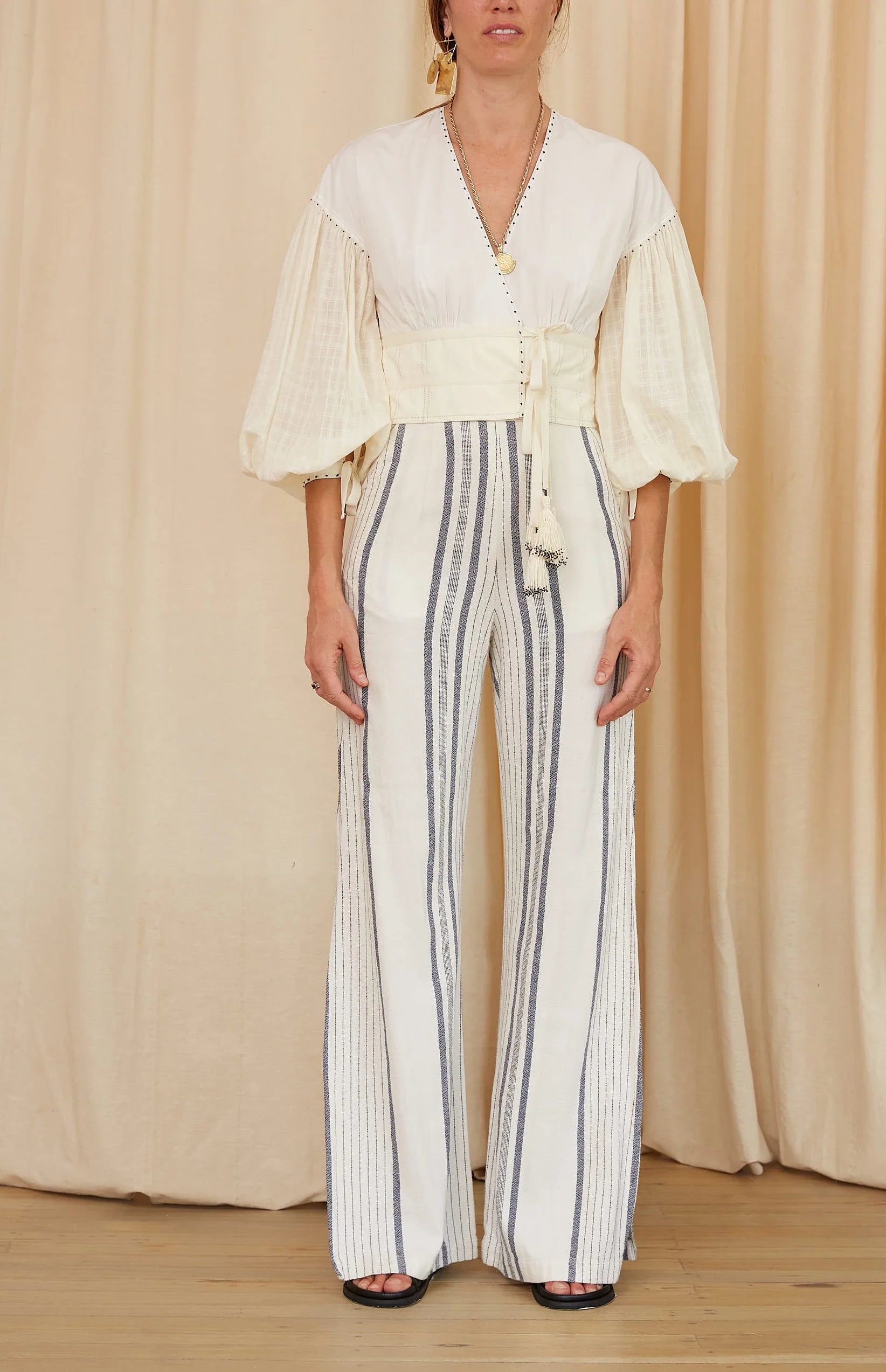 Person wearing a white blouse with bell sleeves and striped pants against a beige curtain background