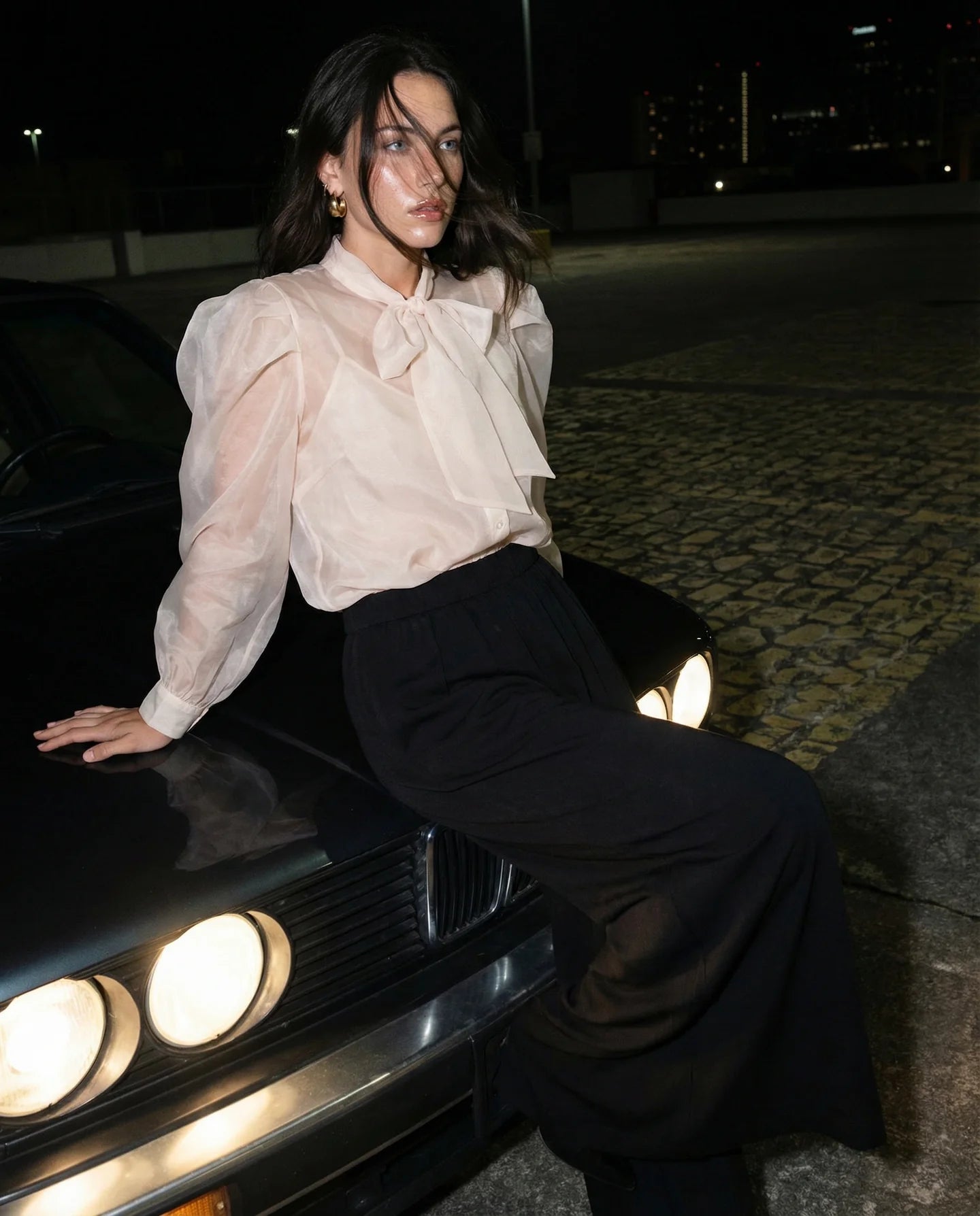 a woman wearing a white blouse and black pants sitting on top of a front of the car