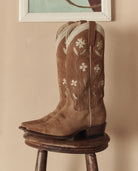 Brown cowboy boots with floral embroidery on a wooden stool against a beige wall.