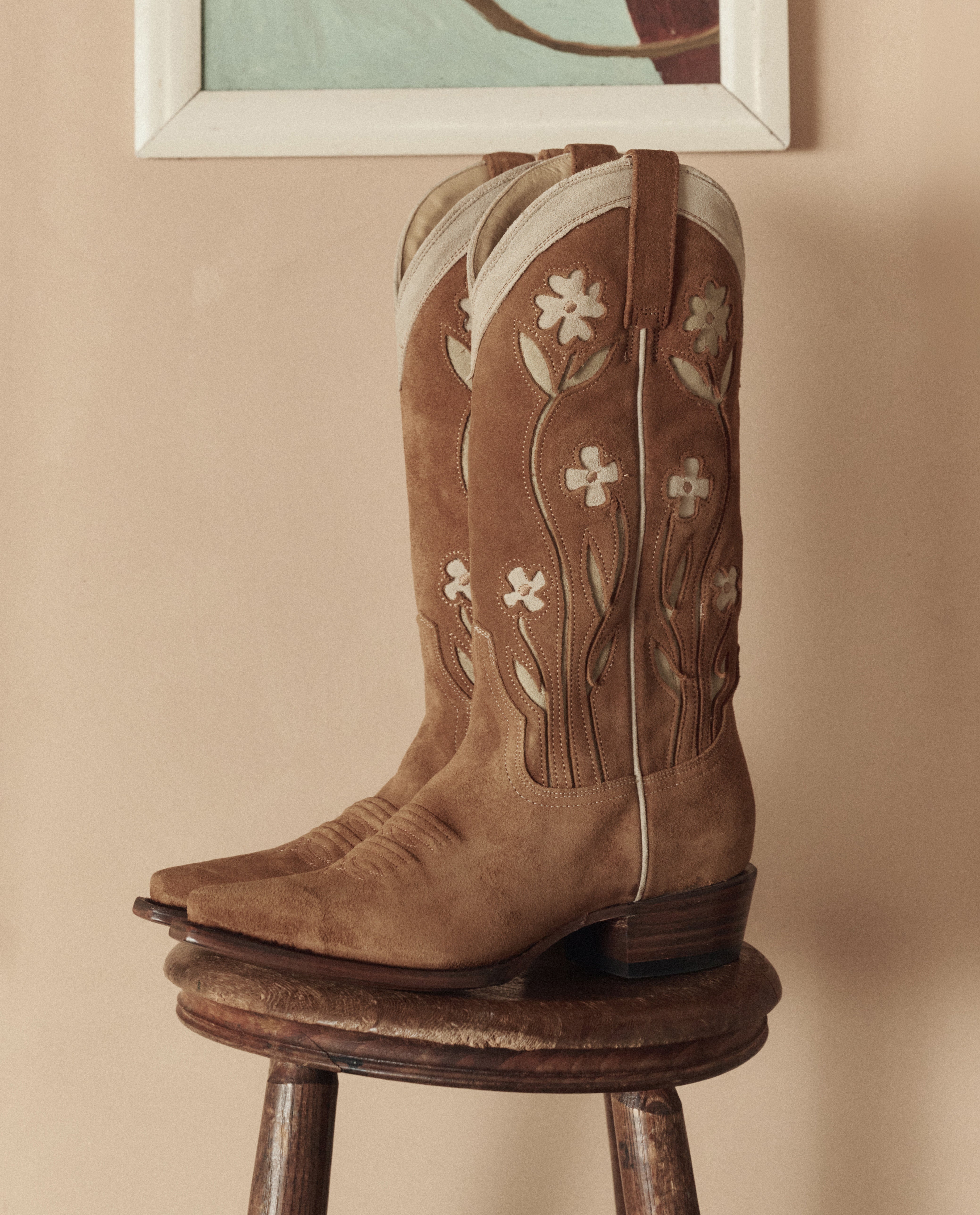 Brown cowboy boots with floral embroidery on a wooden stool against a beige wall.