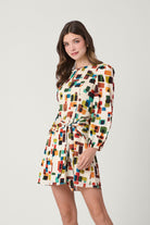 A woman modeling a long-sleeve mini dress with a colorful geometric block pattern on a cream background, set against a plain light gray studio backdrop.