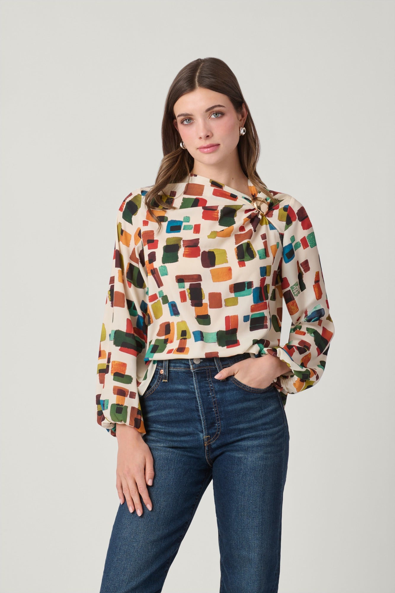 A woman modeling a long-sleeve blouse with a colorful geometric block pattern on a cream background, paired with dark blue jeans, against a plain light gray studio background.