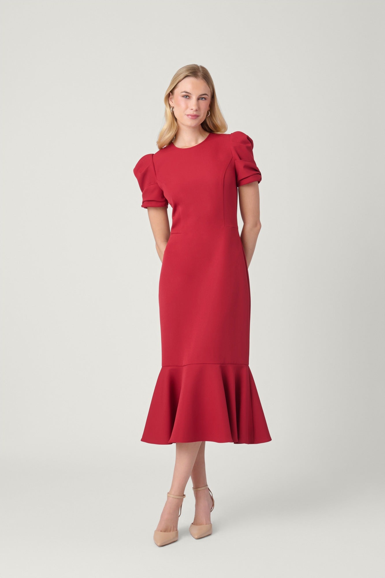 A woman modeling a red midi dress with short puff sleeves and a flared ruffle hem, photographed against a plain light gray studio background.
