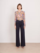 A model stands against a plain off-white wall wearing a long-sleeve button-up blouse with a dense multicolor floral and paisley print, paired with dark wide-leg jeans and black sandals.