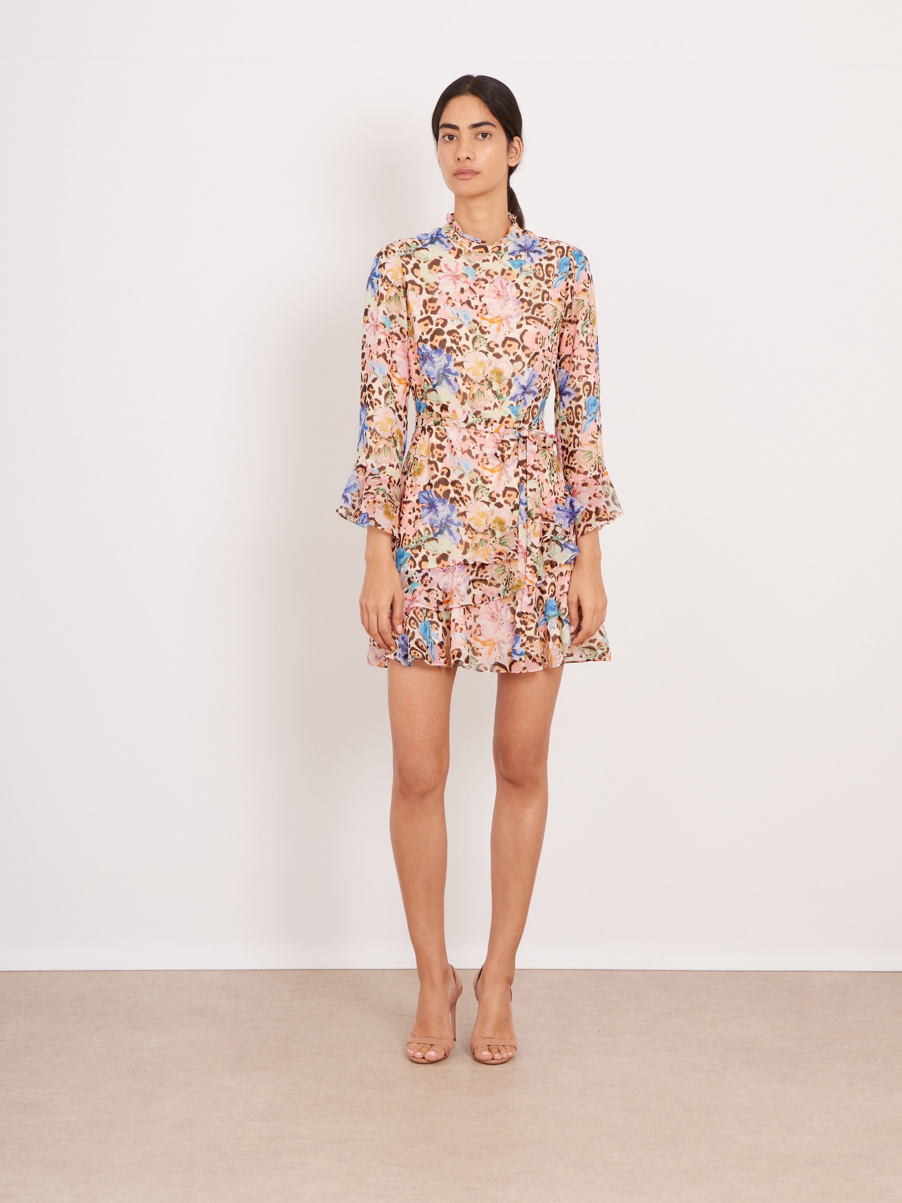 A woman stands against a plain off-white wall wearing a long-sleeve mini dress with a dense pastel floral print in pink, blue, yellow, and green on a light beige base, paired with clear high-heel sandals.