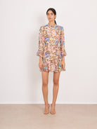 A woman stands against a plain off-white wall wearing a long-sleeve mini dress with a dense pastel floral print in pink, blue, yellow, and green on a light beige base, paired with clear high-heel sandals.