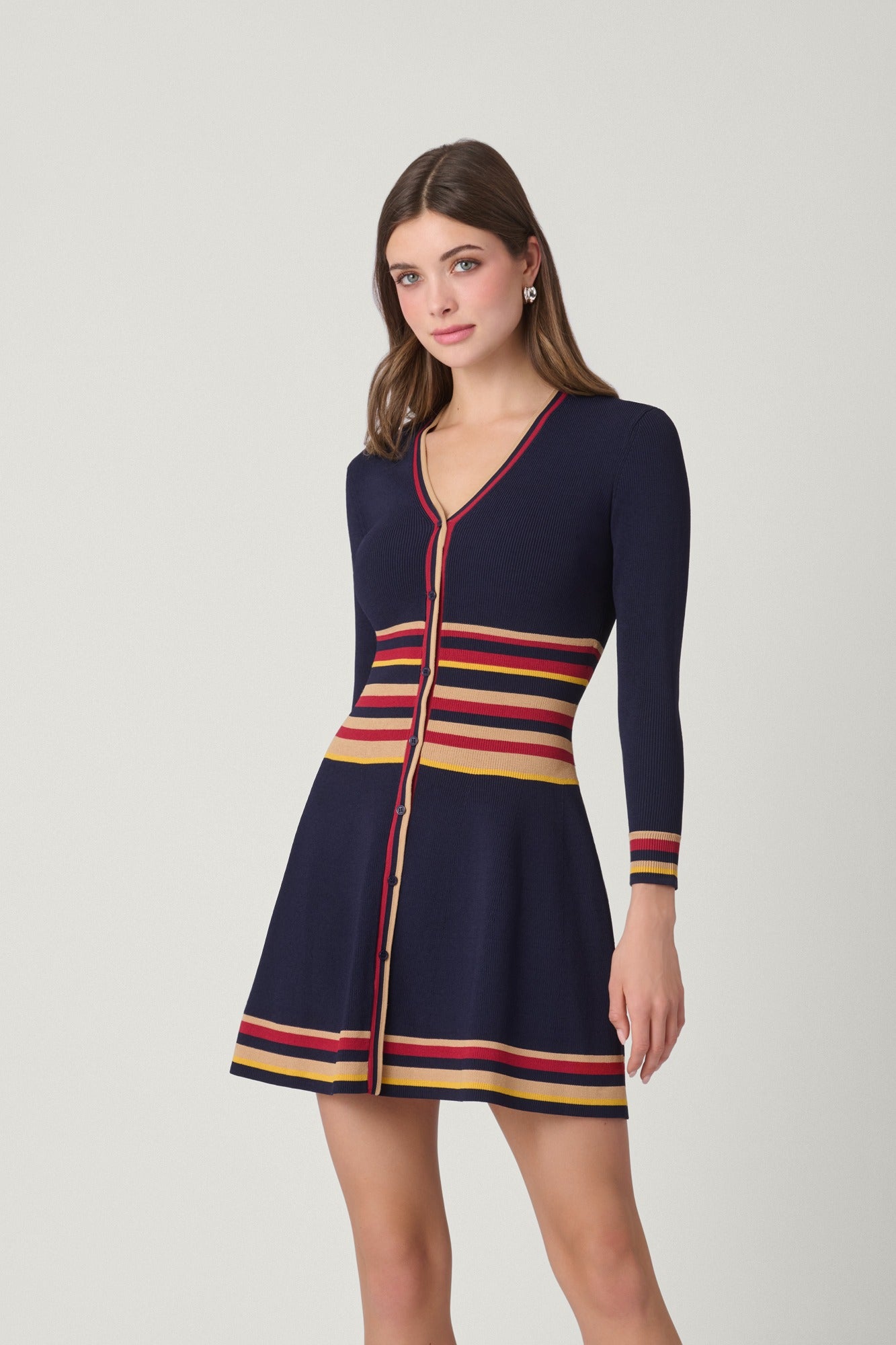 A woman modeling a navy blue knit cardigan dress with red, yellow, and beige horizontal stripes at the V-neck, waist, cuffs, and hem, set against a plain light gray studio background.