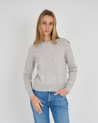 A woman wearing a light gray knit sweater with shoulder cutouts and blue jeans, standing against a light gray studio background.