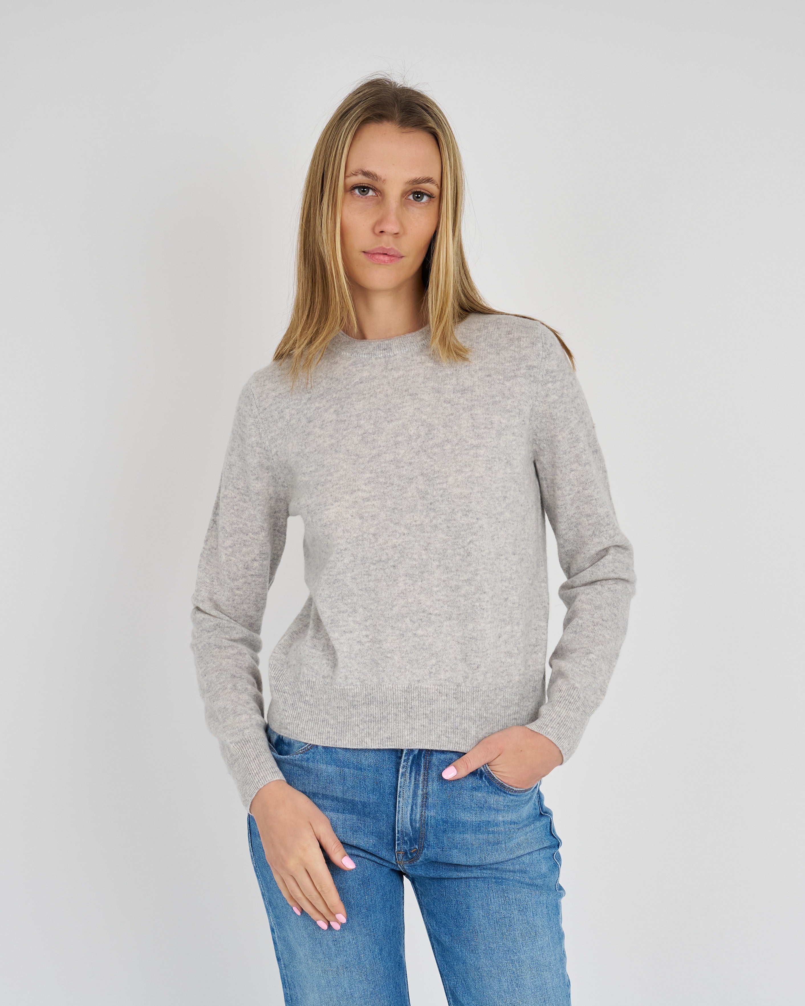 A woman wearing a light gray knit sweater with shoulder cutouts and blue jeans, standing against a light gray studio background.