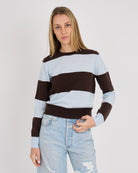 A woman wearing a brown and light blue horizontally striped knit sweater with light-wash distressed jeans, standing against a plain light gray studio background.