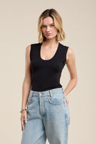 A woman modeling a black sleeveless scoop-neck tank top paired with light-wash blue jeans, set against a solid beige studio background.