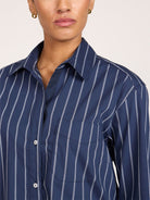 A close up of a model wearing a navy blue and white vertically striped cropped button-down shirt shown from the neck down against a plain light background.