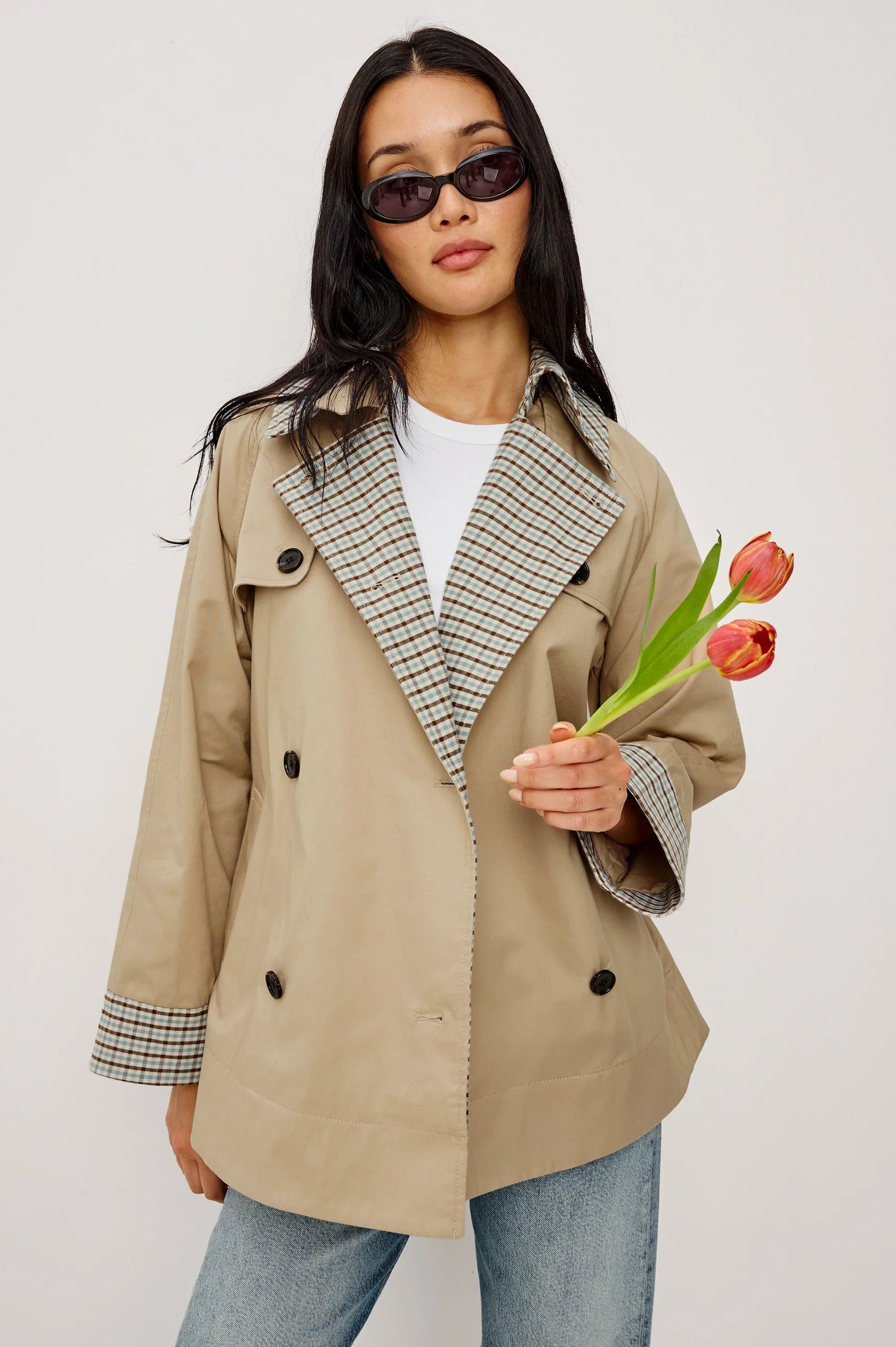 Woman wearing a beige trench coat with plaid accents, holding a tulip against a white background