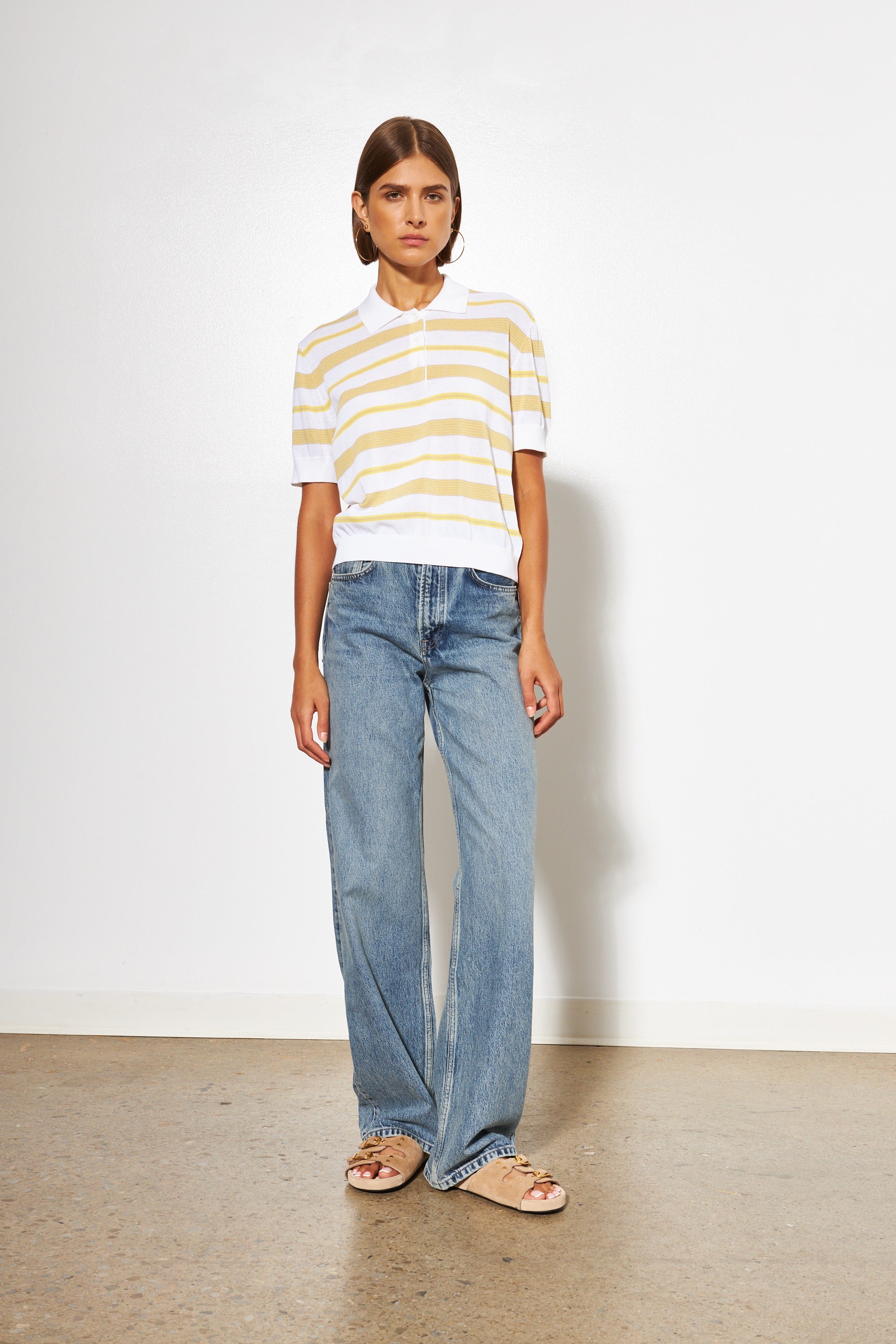 Person wearing a striped shirt and jeans standing against a white wall.